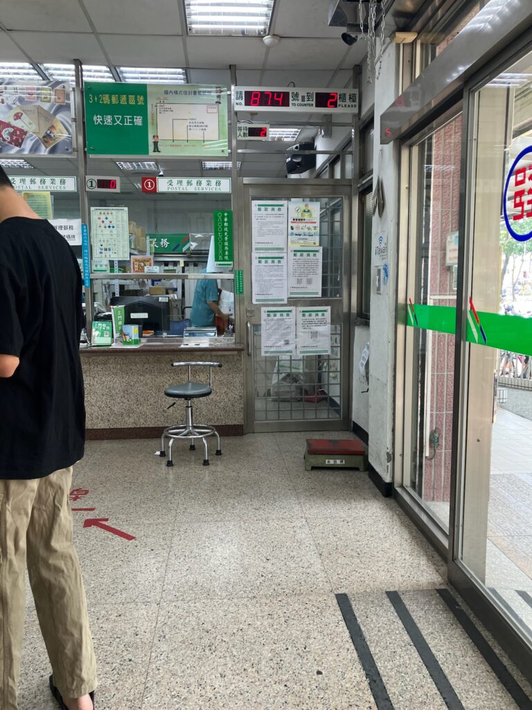 台湾の郵便局内部。電光掲示板に「874号→2番窓口」と表示されており、「受理郵務業務(POSTAL SERVICES)」と書かれた窓口で職員が対応中。待合スペースと丸椅子が見える。