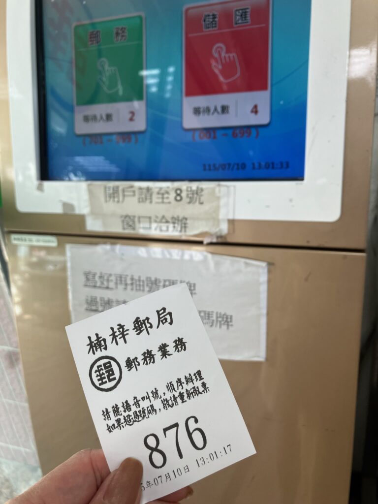 台湾の郵便局(楠梓郵局)の番号券発行機と、取得した整理券。券には「郵務業務・876番」と印字されており、発券時刻は13:01:17。画面には郵務(緑)と貯匯(赤)の2種類のボタンと待ち人数が表示されている。