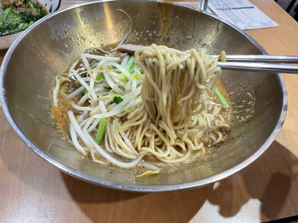 高雄の阿邊麵食(A-Bian Noodles)で提供される台湾乾麺。銀色のボウルに縮れ麺ともやし、青梗菜が盛られ、金属製箸で麺を持ち上げた瞬間